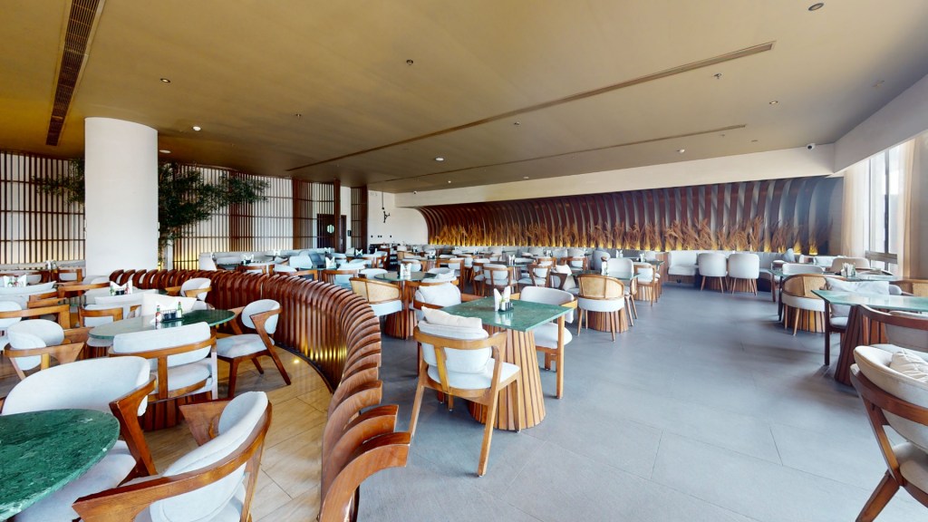 Interior of a modern restaurant featuring wooden furniture, round tables with green marble tops, and soft seating, illuminated by natural light from large windows.