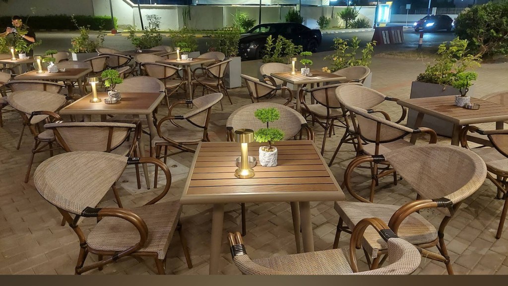 Outdoor dining area with wooden tables and woven chairs, featuring small potted plants and decorative lights.