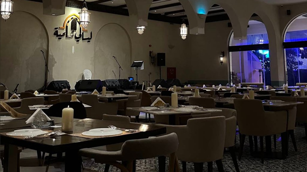Interior of a restaurant featuring elegantly arranged tables, chairs, and decorative lighting, with a stage area and a backdrop displaying the restaurant's name in Arabic.