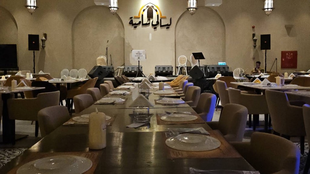 Interior view of a restaurant with long dining tables, set with plates and utensils. Arched walls feature decorative elements, and a stage can be seen in the background. The ambiance is cozy and well-lit.