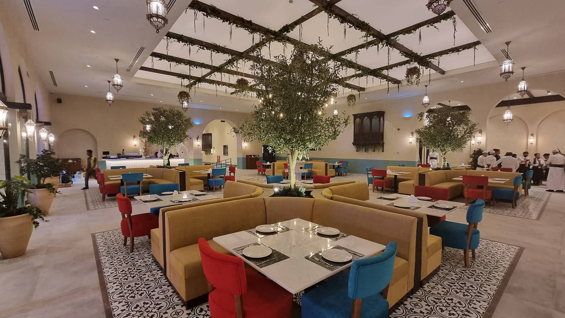 Interior view of a restaurant featuring modern furnishings with colorful seating, large trees, and decorative lighting.