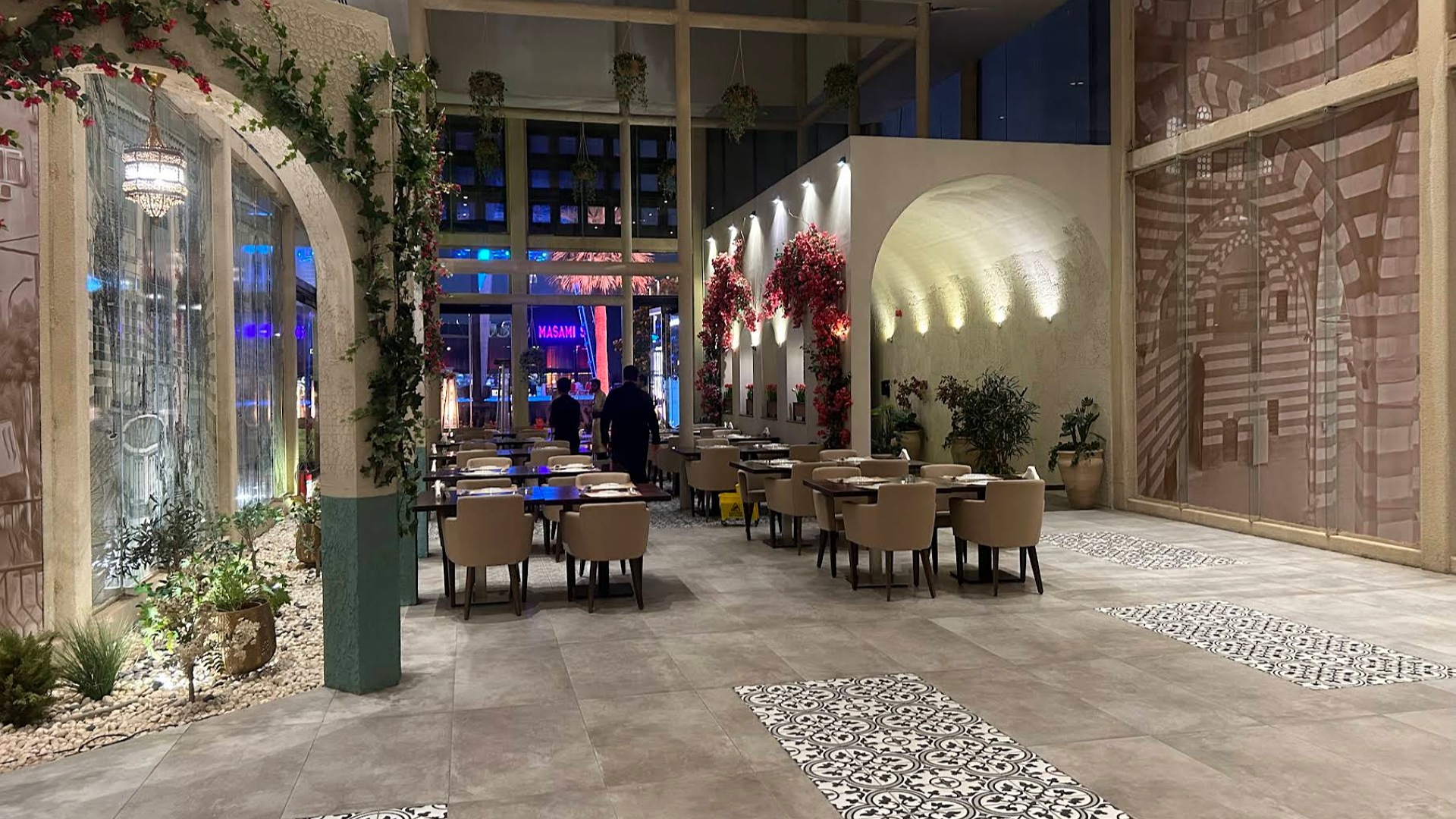 Interior of a stylish restaurant featuring elegant wooden tables, comfortable chairs, and decorative plants. The space has a modern yet cozy ambiance with decorative arches and vibrant floral arrangements.