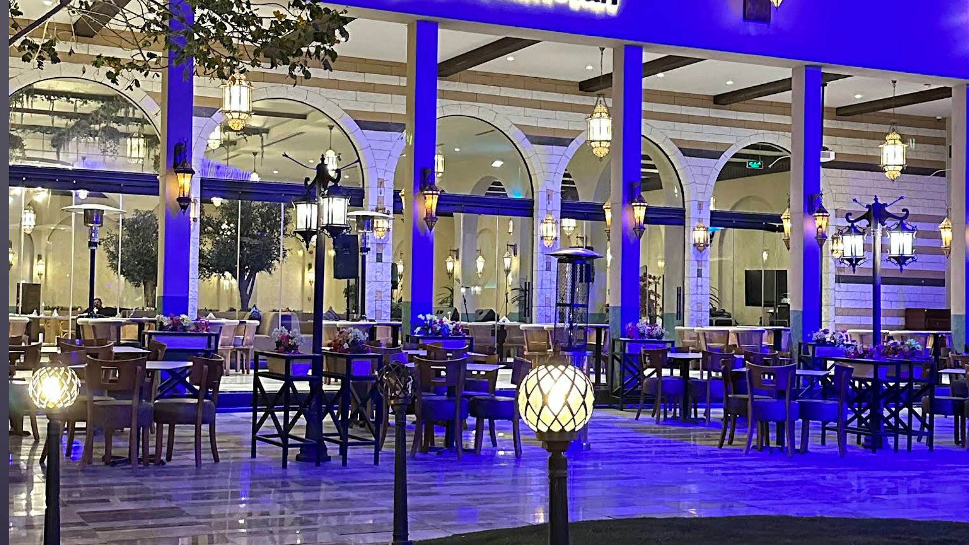 Illuminated outdoor dining area with blue lighting, featuring wooden tables and chairs, decorative lanterns, and potted plants in a stylish restaurant setting.